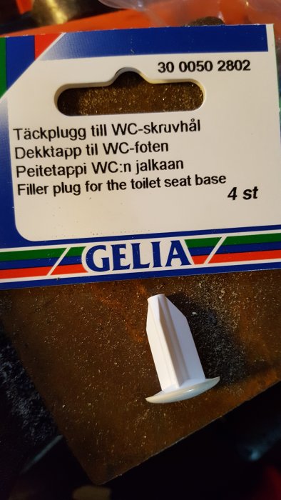 Förpackning och ett exemplar av täckplugg till WC-skruvhål på en brun yta.