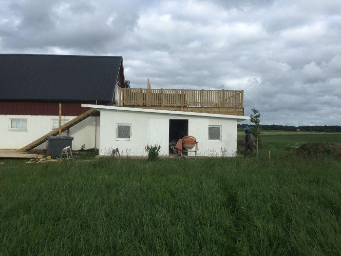 Trädäck med ofärdigt staket ovanpå vit byggnad, arbetsmaterial på marken, öppen landsvy i bakgrunden.