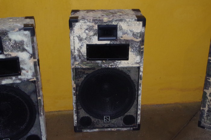 Två högtalare med svart konstlädersklädsel och vit mögelsvamp mot en gul vägg.