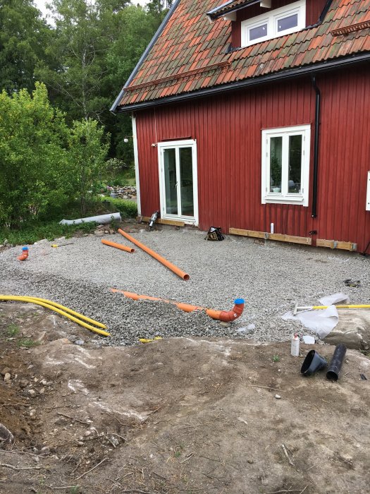 Nydraget dagvattensystem med orange och gula rör synligt ovanför grus på en byggarbetsplats vid en röd stuga.