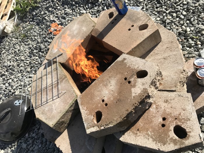 Improviserad tillfällig grill byggd av betongblock med öppen eld och grillgaller, omgiven av grus och ölburkar.