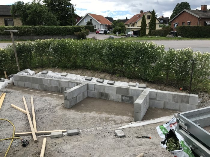 Nybyggd mur av betongblock i en trädgård med byggmaterial runtomkring.