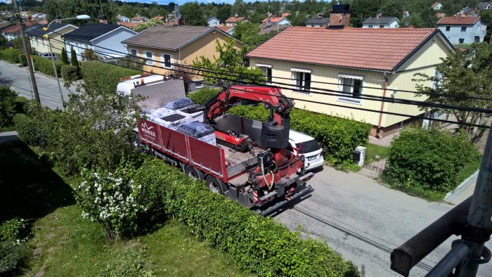 Lastbil med kran lyfter takpannor, arbetsavbrott pga regn, hus och träd i bakgrunden.