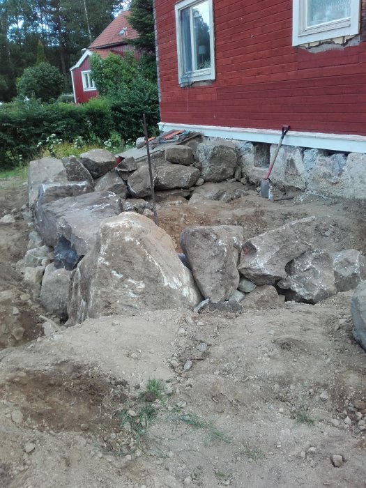 Grävarbete vid husgrund med stora stenblock och grusspade i förgrunden.