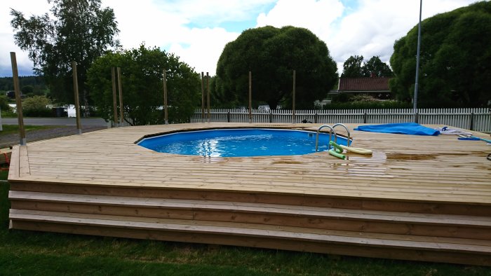 Nybyggd träaltan runt en rund ovanmarkspool i en trädgård med molnig himmel i bakgrunden.