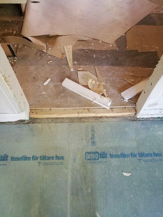 Kuva rikkoontuneesta lattiamateriaalista, jossa näkyy muovinen kosteussuoja, irtonaisia rakennusmateriaaleja ja tuki alla olevan lattian päällä.