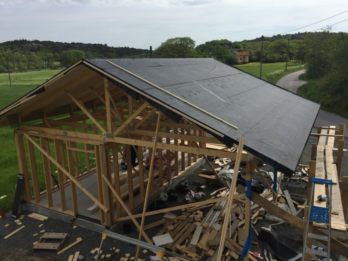 Nybyggt trästomme för hus med mörk takduk delvis lagd på råspontat tak i lantlig miljö.