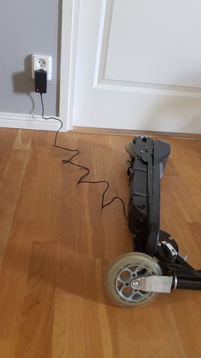 Svart e-scooter liggandes på trägolv ansluten till laddare i vägguttag.