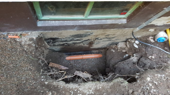 Dräneringsrör för högt i grävd grop nära husvägg med fuktproblem, grus och rötter synliga.