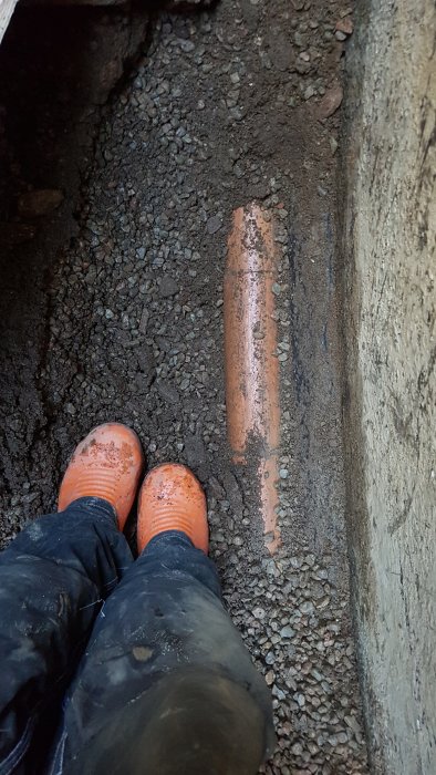 Person i grävd grop med exponerat dräneringsrör och uppfuktad mark.