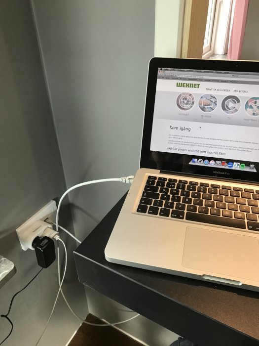 Laptop på svart skrivbord med öppen webbsida, bredvid ett fönster, kopplad till eluttag.