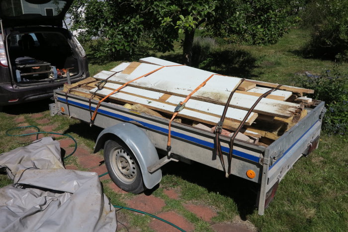 Släpvagn lastad med gamla vitmålade träplankor och takfotsdelar bundna med orange spännband, redo för transport till tippen.