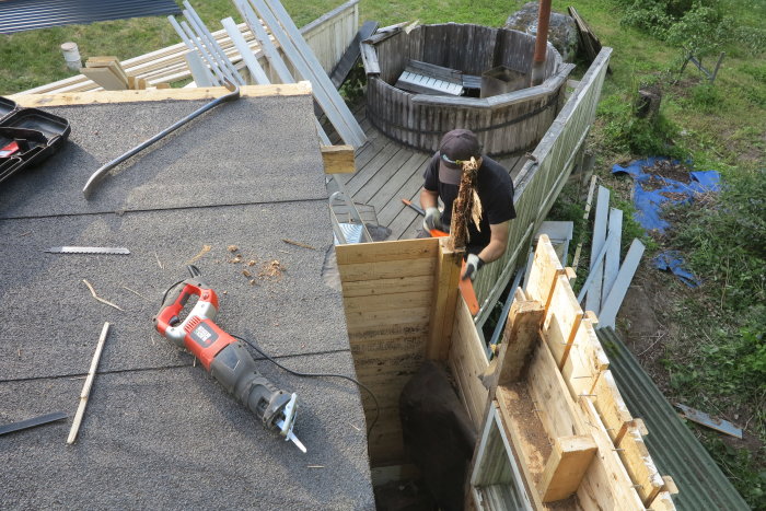 Person reparerar tak med nya träsparrar, gammalt material och verktyg syns på taket.