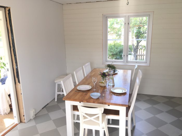 Nyligen renoverat ljust kök med träbord klar för middag, gråvit rutigt golv och väggpanel.