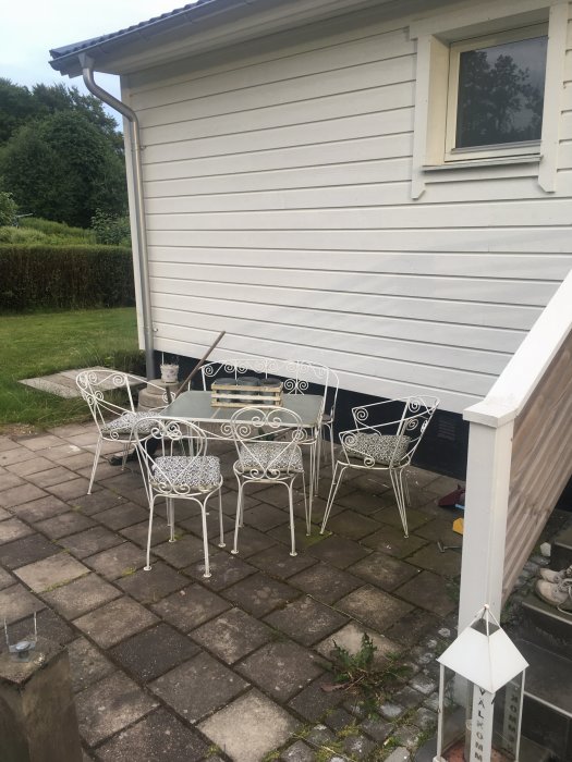 Altan med vitmålat cafebord och stolar framför hus, nära entrétrappa och källartrappa.