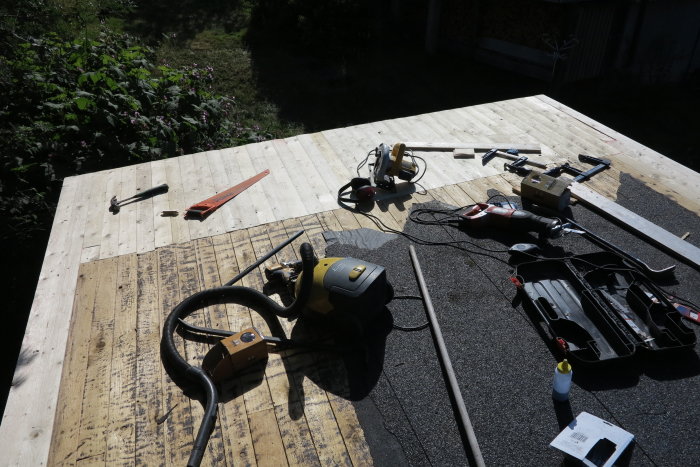 Ny råspont och verktyg spridda på ett tak under renovering, med grönska i bakgrunden.