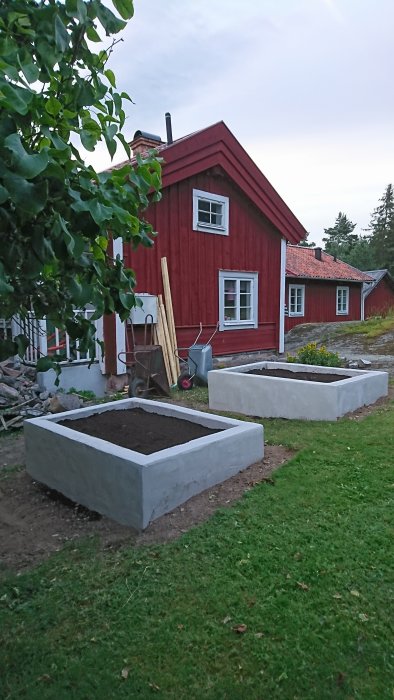 Rödfärgat hus med vita knutar vid nyanlagda högkantade odlingsbäddar fyllda med jord.