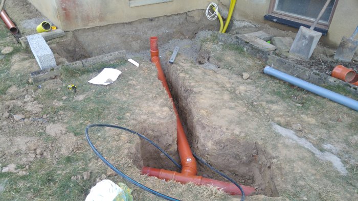 Utgrävning för fundament med synliga rörsystem och verktyg vid en husgrund under installation av värmepump.