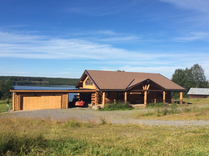 Timrad stuga med anslutande garage framför skog och sjö under klarblå himmel.