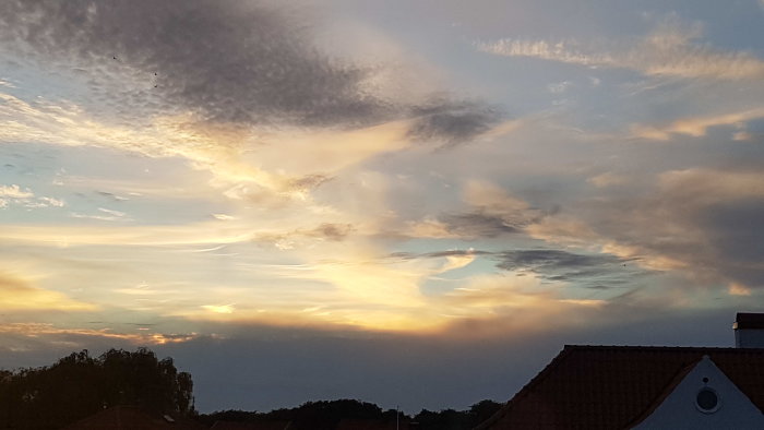 Kvällshimmel med mångfärgade moln över hustak mot solnedgång.