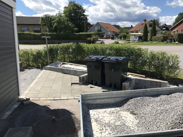 Delvis lagda plattor vid odlingslåda och hög av makadam i ett bostadsområde under konstruktion.