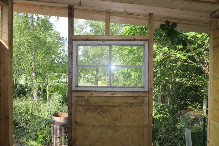 Delvis renoverad gavelvägg med ett fönster med utsikt över grönska, exponering av isolering och reglar.