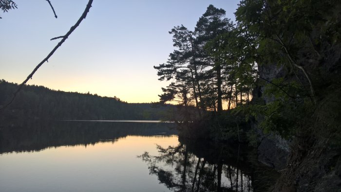 Solnedgång över en stilla sjö i Håverud, Dalsland, med silhuetter av träd och reflektioner i vattnet.