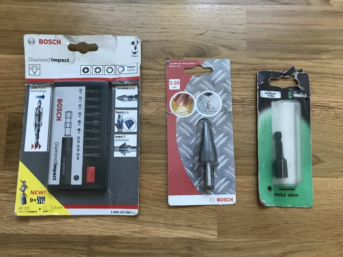 Tre nya förpackningar med verktygstillbehör; Bosch bits set, Bosch stegborr och Wera bits, på trägolv.