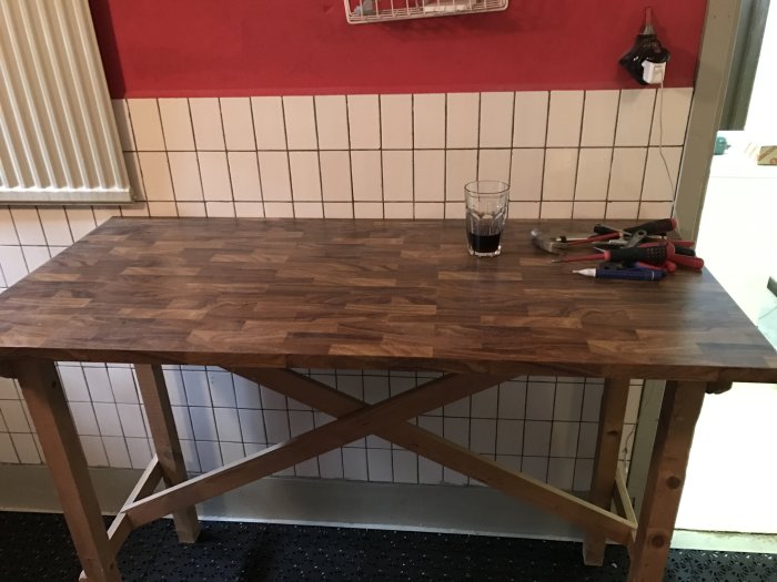 Renoverad arbetsbänk i tvättstuga med träskiva på gammalt bord, verktyg och ett glas på bänken, kakelvägg i bakgrunden.