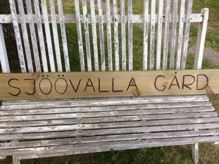 Träskylt med texten "SJÖÖVALLA GÅRD" framför en vit träbänk.