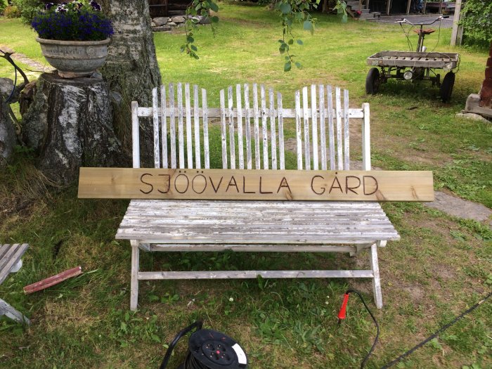Träskylt med texten "SJÖÖVALLA GÅRD" framför en vitmålad bänk med en blomkruka och skottkärra i bakgrunden.