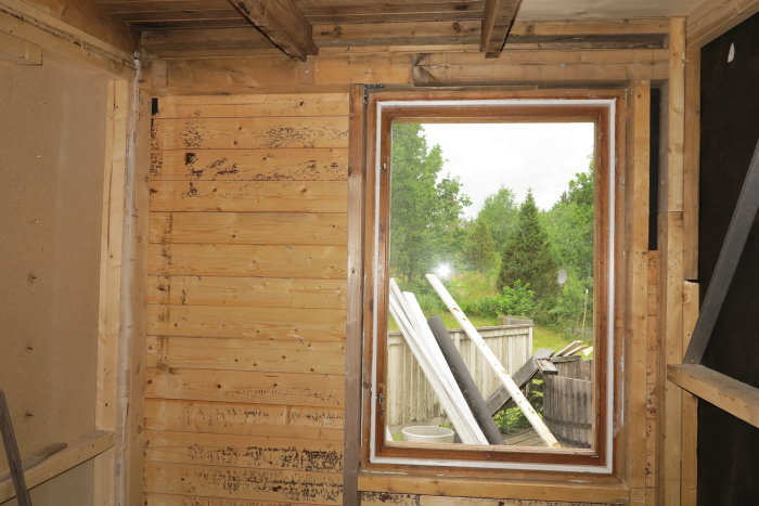 Nytt fönster inpassat mellan trävägg och brandvägg i ett pågående renoveringsprojekt, med utsikt över trädgård.