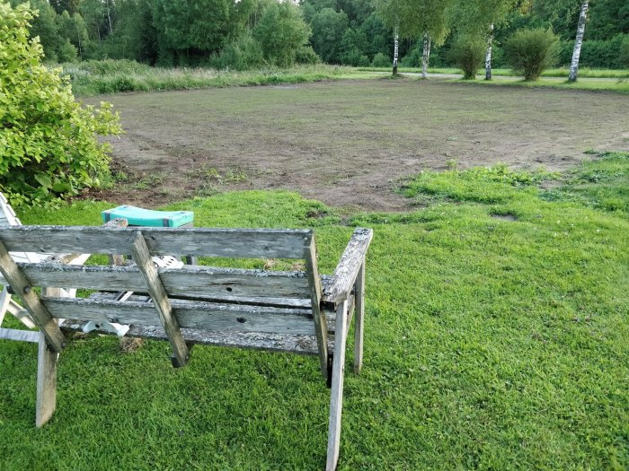 Gammal träbänk med utsikt över en halvt gräsmatta, halvt bar jordyta bakom huset.