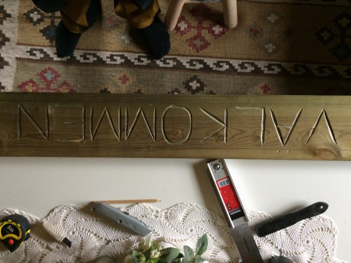 Träplanka med inbränd text "NEW HOME" omgiven av verktyg och en matta i bakgrunden.