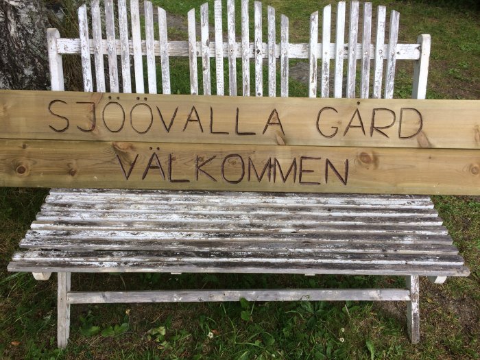 Träskylt med texten "Sjöövalla Gård Välkommen" bränd ovanför en vit sliten trädgårdsbänk.