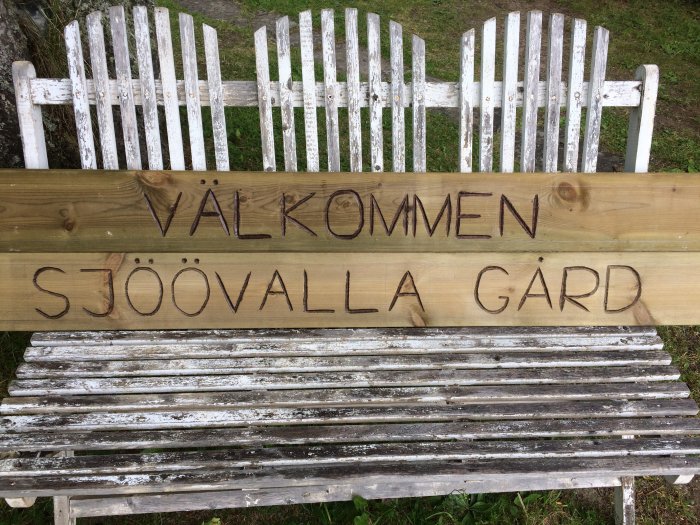 En träskylt med texten "VÄLKOMMEN SJÖOVALLA GÅRD" ingraverad ovanför en sliten vit trädgårdsbänk.