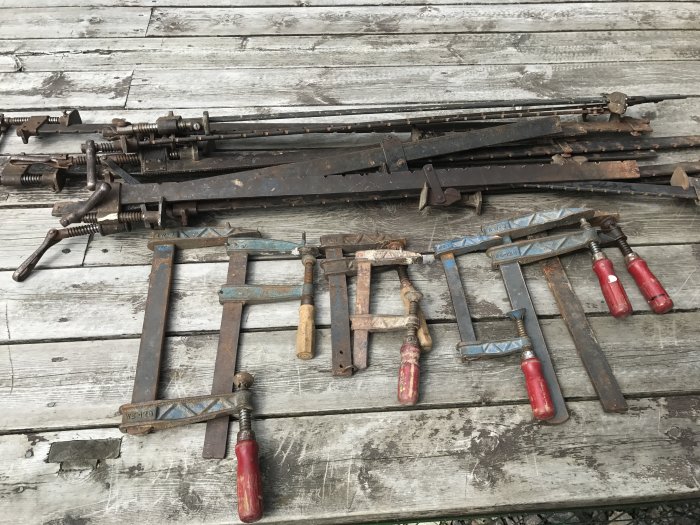 Begagnade verktyg på träbänk, inklusive tvingar och tre skruvtvingar med röda handtag.