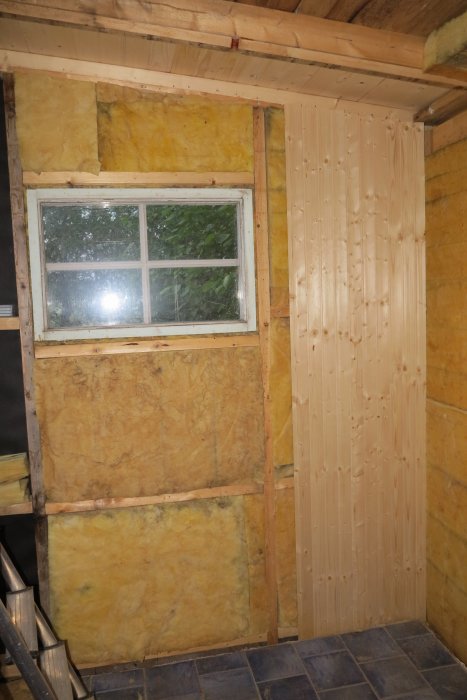En delvis renoverad vägg med ny spikad träpanel och isolering synlig runt ett gammalt fönster.