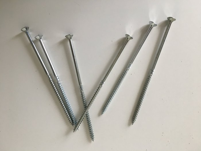 Six long metal screws of varying lengths laid out on a light surface.