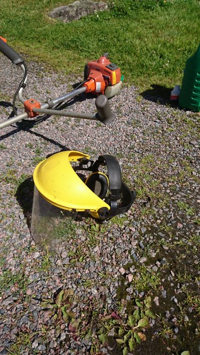 Gult skyddshjälm med visir på grus, bredvid röd grästrimmer i solljus.