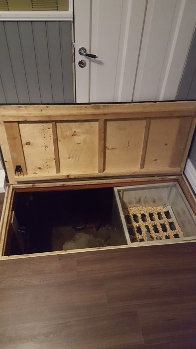 Öppnad golvlucka i hall med gångjärn och vinylklickgolv, visande en nästan osynlig övergång mellan golv och lucka.