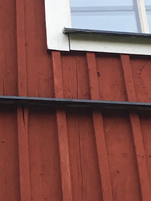 Närbild på nymålad fasad med sprickor i panel och lockläkt, ej skrapade fönsterfoder ovanför.