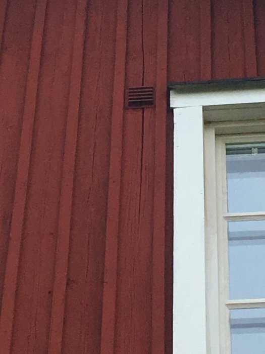 Nymålad husfasad i rött med synliga sprickor på enkel panel och oskrapade fönsterfoder.