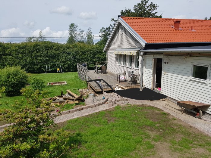 Vit villa med orange tak under renovering med trädgård, gräsmatta, gungställning och påbörjad uteplats.