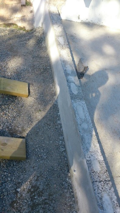 Betongfylld leca-mur vid grusig mark, nära två träbitar. Skugga av person syns till vänster.