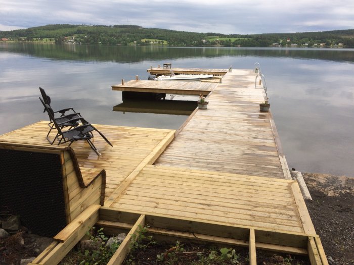 Nybyggd träbrygga som leder ut i en lugn sjö med en fällstol på strandkanten och krukor på bryggan.