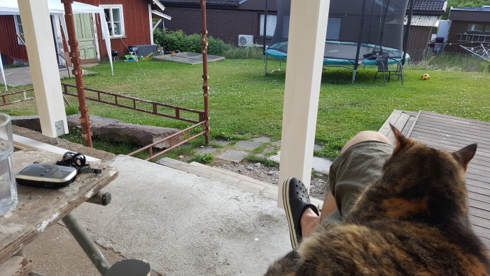 Person med katt i knät på verandan, utsikt mot trädgården med lekstuga och studsmatta.