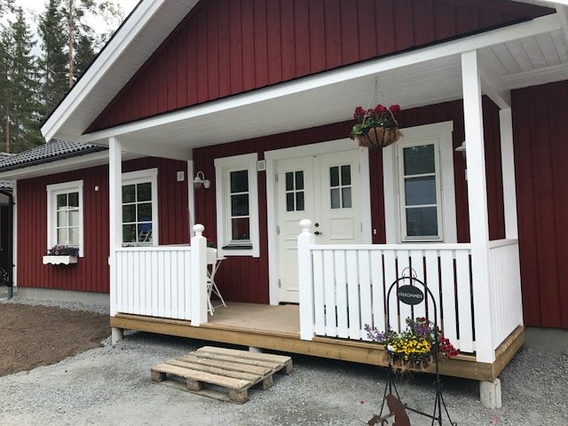 Rödmålat hus med vit veranda, blomlådor, en pall som trappa och grusad uppfart.