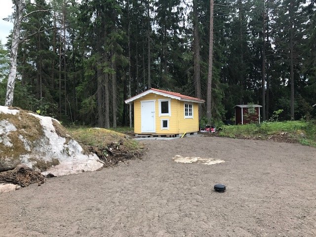 Nyrenoverat gult och vitt litet hus omgivet av nylagd dränering och jord på en trädbevuxen tomt.