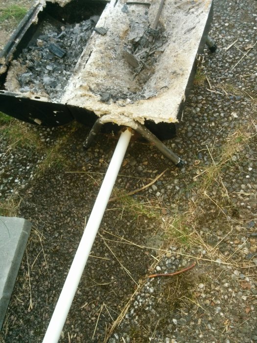 Skadad betongblandare med brända rester och betong, närbild på grusig mark.
